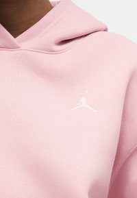 Κοντινό πλάνο ενός ατόμου που φοράει ροζ ανοιχτό hoodie με μικρό λευκό κεντημένο λογότυπο Jumpman στο στήθος.