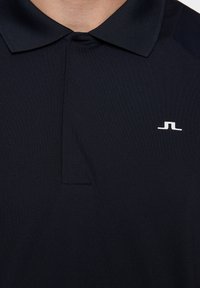 Svart pikétröja av slät tyg med ribbad krage, med en vit logotyp på vänster bröstområde och en standardknäppning.