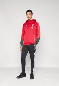 Červená mikina s kapucí s šedými detaily a logem Liverpool FC, doplněná černými kalhotami s bílými bočními pruhy a černými sportovními botami.