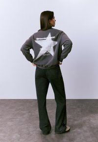 Sudadera gris con un diseño de estrella blanca y texto en la parte posterior; combinada con pantalones vaqueros de pierna ancha negros adornados con patrones de estrellas.