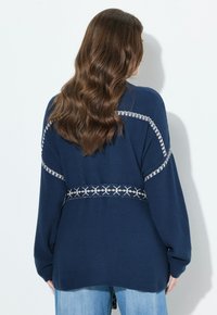 Marinblå stickad cardigan med ett strukturerat mönster, långa ärmar och en kontrasterande vit design längs axlarna och midjan.