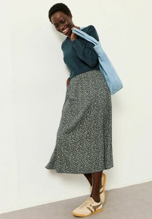 Lächelnde Frau mit dunkelgrünem Pullover mit bestickten Ärmeln, floralem Midirock, beigen Turnschuhen und einer hellblauen Umhängetasche.