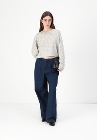 Maglione corto grigio chiaro con un motivo texturizzato, abbinato a jeans scuri a gamba larga. La modella tiene una piccola borsa nera.