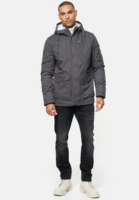 Donkergrijze geïsoleerde jas met capuchon, voorzien van twee voorzakken, houten toggles en een zachte voering. Gemaakt over een donkere spijkerbroek en lichte laarzen.
