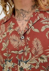 Camisa roja de manga corta con un patrón floral en beige y verde, con collar puntiagudo y cierre de un solo botón.