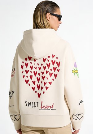 Femme portant un sweat à capuche beige avec un motif de cœur rouge et le texte "SWEET heart" dans le dos, lunettes de soleil noires et pantalon beige.