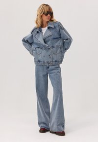 Kobieta woversize'owej dżinsowej kurtce, z rozszerzanymi spodniami dżinsowymi, ciemnymi okularami przeciwsłonecznymi i brązowymi butami, stojąca na tle jasnej, jednolitej ściany.