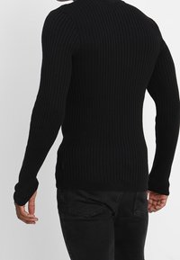 Svart ribbad stickad tröja med långa ärmar, åtsittande design, rundad halsringning och sidslitsdetaljer, bärs över svarta jeans.