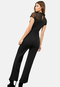 Frau in einem schwarzen Jumpsuit mit transparenten kurzen Ärmeln und einem Schlüssellochdetail am Rücken, steht mit leicht gedrehtem Rücken.