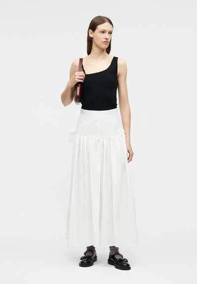 Γυναίκα που φοράει μαύρο τοπ με έναν ώμο, λευκή μακριά φούστα με ψηλή μέση, γκρι κάλτσες, μαύρα μοκασίνια και κρατά καφέ τσάντα ώμου.