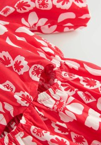 Rode stoffen jurk met een witte bloemenprint. Het heeft elastische plooien bij de halslijn en de mouwen voor een comfortabele pasvorm.