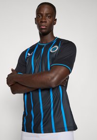 Homem em pé com os braços cruzados, vestindo uma camisola de futebol do Club Brugge, listrada em preto e azul, e calções brancos, contra um fundo simples.