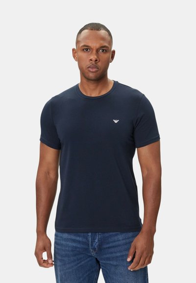 T-paita, jossa on lyhyet hihat, tummansininen, sileää puuvillaa, pyöreä kaula-aukko ja minimaalinen logotyyppi rinnassa. Yhdistetty sinisiin farkkuihin.