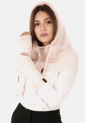 Giovane donna che indossa una sciarpa a cappuccio lavorata a maglia rosa e guanti senza dita abbinati, vestita con una camicia bianca e pantaloni neri, in posa su uno sfondo bianco.