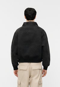 Άνδρας που φοράει μαύρο bomber μπουφάν και μπεζ παντελόνι cargo, φωτογραφημένος από πίσω με φόντο απλή ανοι χτή επιφάνεια.