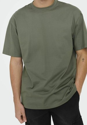 Mężczyzna w jednolitą oliwkową koszulkę z krótkim rękawem i okrągłym dekoltem oraz czarne spodnie, z prawą ręką w kieszeni i małym tatuażem krzyża na lewym ramieniu.