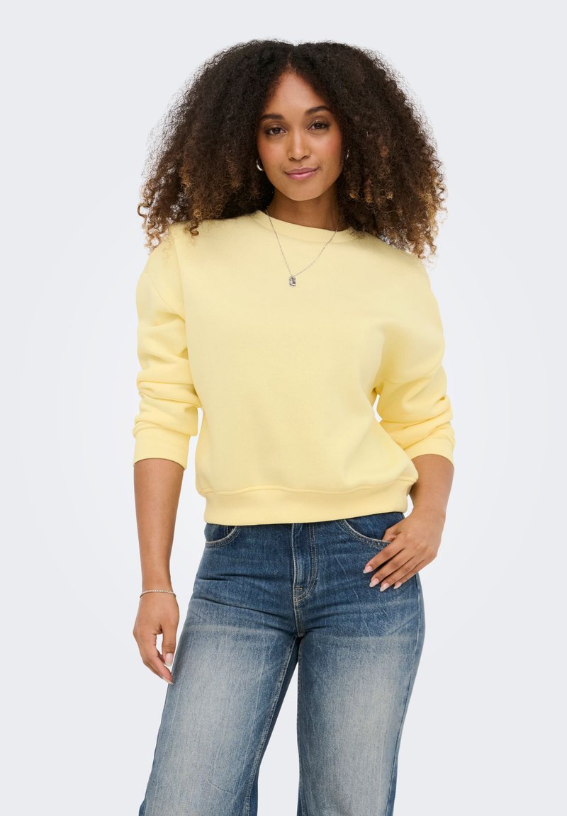 Femme aux cheveux bouclés portant un sweat-shirt jaune clair et un jean bleu, debout avec une main sur la hanche devant un fond uni.
