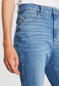 Närbild av en person som bär ljusblå denimjeans med en liten "BOSS"-etikett på framsidan av fickan och en avspänd hand vid sidan.