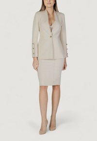 Blazer ajustado beige con mangas largas y detalles de botones dorados, combinado con una falda lápiz ceñida. Tela suave y diseño elegante.