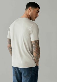 T-shirt beige chiaro a maniche corte con collo rotondo, texture liscia e vestibilità rilassata, abbinato a jeans blu scuro. Tatuaggi visibili sul braccio.