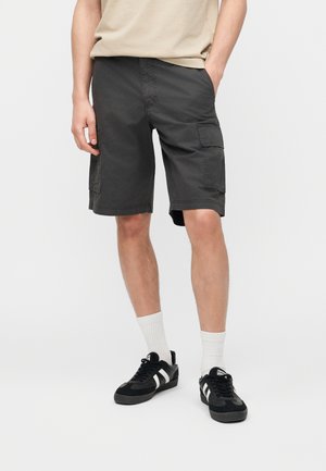 Muškarac u tamnosivim cargo kratkim hlačama, bež majici, bijelim čarapama i crnim tenisicama s bijelim prugama, stoji sa rukama u džepovima.