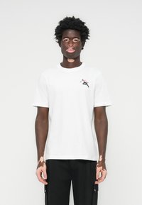 Weißes Baumwoll-T-Shirt mit kurzen Ärmeln, Rundhalsausschnitt, mit einem kleinen grafischen Motiv einer springenden Figur in Rot und Schwarz auf der Brust.