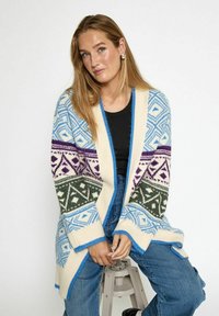 Cardigan lavorato a maglia multicolore con motivi geometrici blu, verde e viola su uno sfondo crema; apertura frontale e polsini a costine.