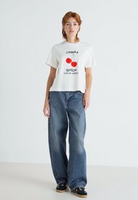 Weißes Baumwoll-T-Shirt mit einem Kirschgrafik und Text; kombiniert mit weit geschnittenen blauen Denim-Jeans; das Model steht vor einem neutralen Hintergrund.