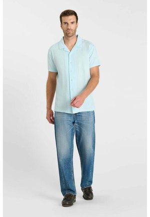 Man draagt lichtblauw overhemd met korte mouwen, blauwe spijkerbroek en zwarte schoenen, staand tegen een effen witte achtergrond.