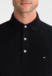 Black polo shirt made from cotton, featuring a classic collar, three white buttons, and a small red, white, and blue logo on the chest.