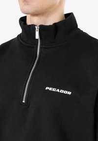 Pullover nero con zip a mezza lunghezza e colletto a costine, dotato di una zip argentata e logo "PEGADOR" ricamato in bianco sul petto. Texture morbida.