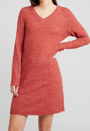 Kobieta w miękkiej, długiej, czerwonej sukience swetrowej z dekoltem w serek, trzymająca jedną rękę na biodrze na jednolitym tle.