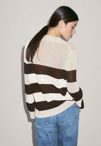 Donna con capelli scuri raccolti in una coda bassa, che indossa un maglione a righe crema e marrone scuro e jeans blu, rivolta di lato contro un muro neutro.