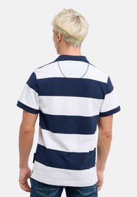 Marinblå och vit randig pikétröja med korta ärmar, ribbad krage och en tagg vid fållen. Tillverkad av bomull med en mjuk textur.