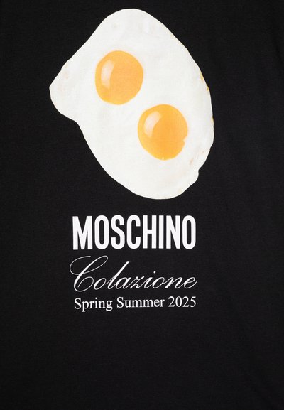 Camiseta negra con un gráfico de un huevo frito con dos yemas amarillas. El texto incluye "Moschino", "Colazione" y "Primavera Verano 2025" en blanco.