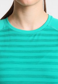 Tealfärgad träningströja med horisontella ränder, rund hals, korta ärmar och ett mjukt, lätt material. Sömmen är synlig längs kanterna.