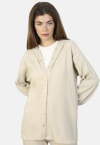 Beige Kapuzenhemd mit Knopfleiste aus strukturiertem Gewebe und langen Ärmeln. Es hat einen geraden Schnitt, sechs Knöpfe und eine lockere Passform.