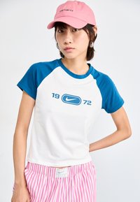 Valge T-särk siniste raglanvarrukatega, millel on sinine Nike logo ja "1972" tekst. Kombineeritud roosade triibuliste lühikestega.