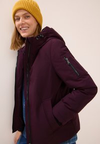 Doudoune violette avec capuche, dotée d'une poche zippée sur la manche et de poches latérales, accompagnée d'un bonnet en tricot jaune.