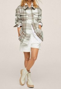 Geruite shirtjack in beige en wit, met twee voorzakken en knoopsluiting, gecombineerd met witte shorts en lichtgekleurde enkellaarzen.