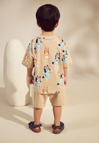 T-shirt en coton beige avec des personnages de chiens dessin animés colorés, associé à un short clair et des sandales foncées, vu de dos.