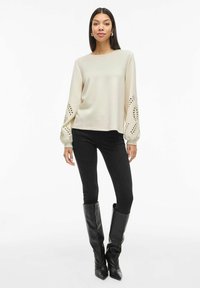 Blusa beige a maniche lunghe con dettagli a taglio sulle maniche, abbinata a jeans skinny neri e stivali neri fino al ginocchio.