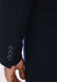 Morski wełniany płaszcz ma rękawy z delikatną teksturą i trzy czarne guziki przy mankietach, co podkreśla dopasowany krój.