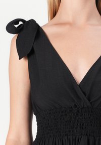 Vestido preto com um decote em V profundo, corpete franzido e laços de tecido nas alças dos ombros. O material tem um acabamento texturizado.