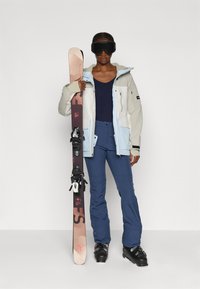 Skisportklær som inkluderer en beige og lyseblå isolert jakke, marineblå topp og blå skibukser. Holdes en brun ski med svarte bindinger.