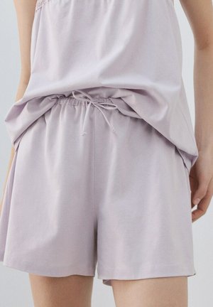 Ženska, oblečena v svetlo vijolične elastične kratke hlače z zavezanim vrvico in ujemajočo ohlapno majico, stoji pred nevtralnim ozadjem.
