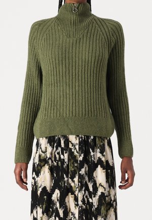 Pull en maille côtelée verte avec col zippé haut, associé à une jupe boutonnée à motif abstrait noir, blanc et vert.