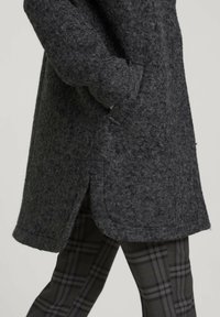 Cappotto in feltro grigio scuro con tasche laterali, orlo arrotondato e leggere spaccature ai lati. Abbinato a pantaloni a quadri neri e grigi.
