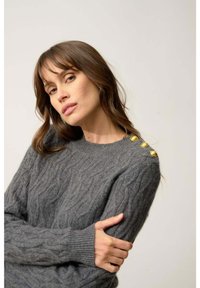 Szary sweter w splot sznurkowy z prążkowanymi mankietami i dekoltem, z trzema złotymi guzikami na lewym ramieniu. Miękka tekstura, luźny krój.