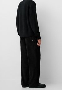 Person wearing loose black corduroy pants, black knit sweater, and black shoes, standing sideways against a plain light background.